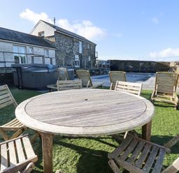 An outdoor patio area with a round wooden table and chairs on grass near a stone building and a swimming pool at Penpethy Farm in Tintagel