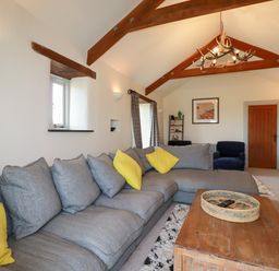 A living room with a grey sectional sofa with yellow cushions a wooden coffee table under wooden ceiling beams at Penpethy Farm in Tintagel