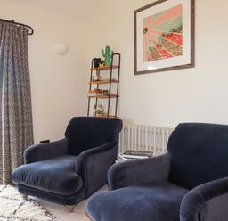 Two dark armchairs near a window with patterned curtains a small shelf with decorative items and a framed picture on the wall at Penpethy Farm in Tintagel
