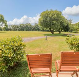 Two wooden chairs on a patio facing a grassy area with trees and a pathway at Lodge 16 in Lanteglos near Camelford
