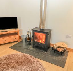 A wood stove with fire burning next to a TV on a wooden stand and firewood on a rack at The Mews in Bovey Tracey near Chudleigh Knighton