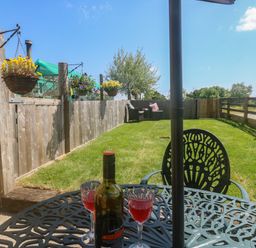 A garden with a metal table and two glasses of red drink on it a bottle of wine hanging flower baskets a wooden fence and chairs at The Mews in Bovey Tracey near Chudleigh Knighton