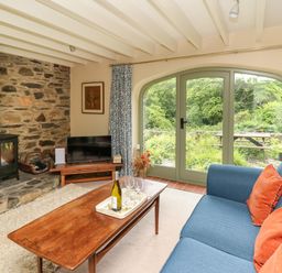 A living room with a wood stove next to a stone wall and large glass doors showing greenery outside at Taibach in Dinas Cross