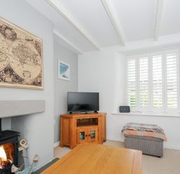 A living room with a wood burning stove a TV on a wooden cabinet a gray ottoman a wooden coffee table and a window with white shutters at Quay House in Charlestown