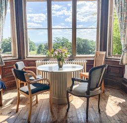 A round table with a flower vase and four chairs near large windows with curtains at Nethercott House in Iddesleigh near Hatherleigh