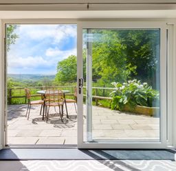 A patio with a round table and four chairs surrounded by plants and trees seen through sliding glass doors at Goose Cottage within the Helland Barton Farm collection in Delabole