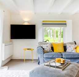 A living room with a blue sofa yellow cushions a wall-mounted TV and a window showing trees at Goose Cottage - within the Helland Barton Farm collection in Delabole