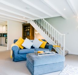 A living room with blue sofa and yellow cushions next to stairs and an open kitchen in the background at Goose Cottage within the Helland Barton Farm collection in Delabole