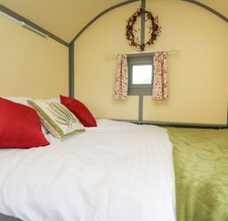 A bedroom with a bed with red and green pillows a small window with floral curtains and a wreath on the wall at Brown Hare Shepherds Hut in Llangorse