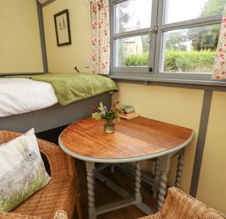A small room with a wooden table holding a vase of flowers and books next to a bed with a green blanket and wicker chair with a rabbit pillow by a window at Brown Hare Shepherds Hut in Llangorse