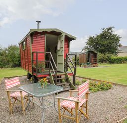 A red gypsy-style caravan with open door on a gravel area with table and two striped chairs at Tilly Gypsy-style Caravan Hut in Llangorse