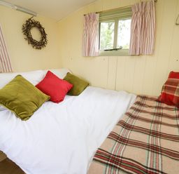 A bed with white sheets green and red pillows a plaid blanket and a window with striped curtains in a small room at Tilly Gypsy-style Caravan Hut in Llangorse