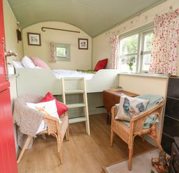 A small room with a red door two wicker chairs a bed with a ladder wooden floor and floral curtains at Sweet Briar Shepherds Hut in Llangorse