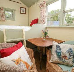 A small room with a raised bed with a small ladder a wooden table with a vase of flowers and two wicker chairs with animal print cushions at Sweet Briar Shepherds Hut in Llangorse