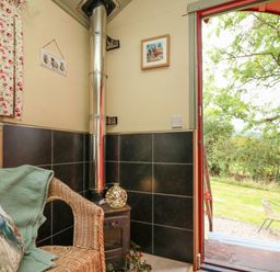 A small seating area with a wicker chair and wood stove next to an open door showing a garden at Sweet Briar Shepherds Hut in Llangorse