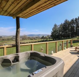 An outdoor decking area with a hot tub a wooden picnic table and a view of fields and hills at Llety Cariad in Llandrindod Wells