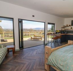 A room with a bed a striped armchair a kitchen area and folding glass doors opening to a wooden deck with a picnic table at Llety Cariad in Llandrindod Wells