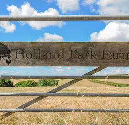 A wooden sign on a metal gate with fields and trees in the background at Holland Park Farm