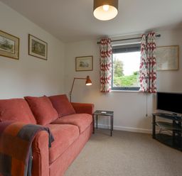 A room with a red sofa and floor lamp next to a window with patterned curtains and a TV on a glass stand at Lillemor South Tawton near Whiddon Down