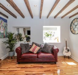 A living room with a red leather sofa and cushions a guitar large wall clock plants wooden beams on ceiling and artwork at O'Neil Cottage in Beadnell