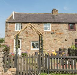 A stone cottage with a black picket fence and seating area with cushions in the garden at O'Neil Cottage in Beadnell