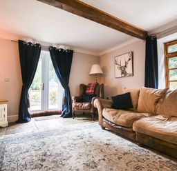 A living room with a beige sofa black cushions an armchair with a lamp near a deer painting a wooden cabinet with a TV blue curtains and a patterned rug at Barley Cottage Weycroft near Axminster