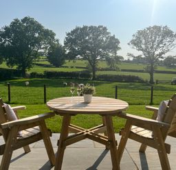 A wooden table with two glasses of drink and a potted plant between two wooden chairs on a patio overlooking a green field with trees and sheep at Yr Onnen in Bontuchel near Ruthin