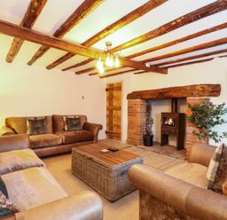 A living room with wooden beams on the ceiling two brown sofas a wooden coffee table a fireplace with a wood-burning stove and a wall-mounted TV at Well Cottage in Grimston