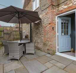 An outdoor patio with a round table and four wicker chairs under a large umbrella next to a stone wall with a blue door at Well Cottage in Grimston
