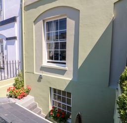 An exterior view of a light green building with two white-framed windows and flowers in pots near the windows at Drake's Retreat in Plymouth