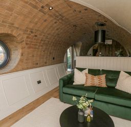 A living area with a green sofa and cushions a round window and a round black table with decorative items at Tan y Wawr- Onnen in Llanfair Caereinion