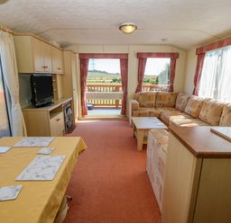 A living area with an L-shaped sofa a coffee table a dining table with placemats a television unit and large windows at Gorphwysfa Caravan in Tregele near Cemaes Bay