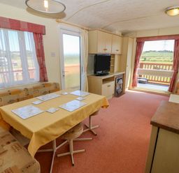 A living area with a dining table, bench seating, television on a cabinet, and windows with red curtains at Gorphwysfa Caravan Tregele near Cemaes Bay