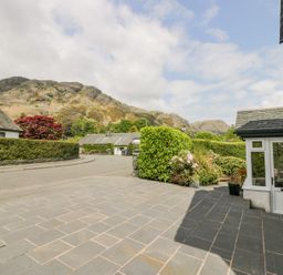 A paved driveway with bushes and flowers next to a white house with mountains in the background at Holme Fell in Coniston