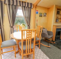 A dining area with a round wooden table and four chairs next to a window with striped curtains and a gray rocking chair beside a fireplace at Rosehaven in Tegryn near Crymych