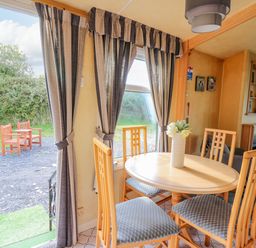 A dining area with a round wooden table and four chairs next to windows with curtains and an open door showing two wooden chairs outside at Rosehaven in Tegryn near Crymych