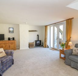 A living room with two armchairs a wooden cabinet a wood stove and glass doors with yellow curtains at Cattle Tree Cottage in Llangoedmor near Cardigan