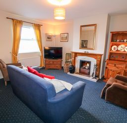 A living room with a blue sofa, brown armchairs, a wooden cabinet with plates and cups, a fireplace with a mirror above it, a television on a wooden stand, and yellow curtains on the window at 17 Buninver Road in Gortin
