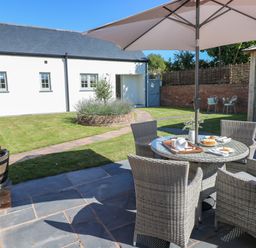 A garden patio with woven chairs around a table under an umbrella with a white building and a grassy area in the background at Catamouse in Milford Haven