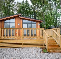 A wooden cabin with a deck and stairs surrounded by trees and gravel at Bluebell Lodge in Gargrave