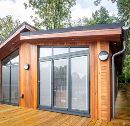 A wooden cabin with large glass doors and windows on a wooden deck surrounded by trees at Bluebell Lodge in Gargrave