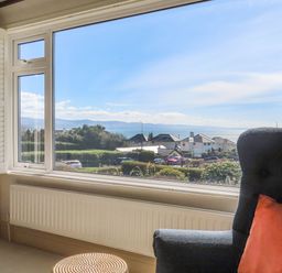 A window with a view of houses trees and water with a chair and cushion by Awelfryn in Criccieth