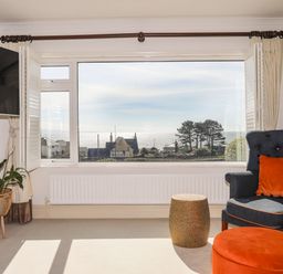 A room with a large window showing a sea view a blue armchair with an orange cushion an orange ottoman a plant in a stand and a mounted television at Awelfryn in Criccieth