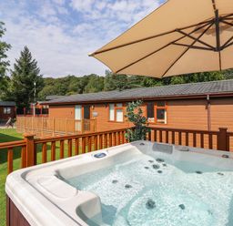 An outdoor hot tub on a wooden deck with an umbrella and a brown wooden cabin in the background at Honeysuckle Lodge in Thirlmere 14 White Cross Bay near Windermere