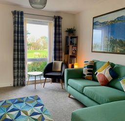 A living room with a green sofa a black armchair a small round table a bookshelf and a large window with curtains at 2 Boathouse Terrace in Mawgan Porth