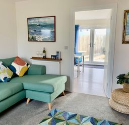 A living room with a green sofa and colorful cushions two paintings on walls and a round wicker side table with a plant at 2 Boathouse Terrace Mawgan Porth