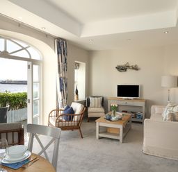 A living room with a sofa armchairs a coffee table and a TV near an arched window with a sea view at 1 Argyle House Lyme Regis