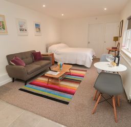 A room with a brown sofa with purple pillows a bed with white bedding a wooden coffee table on a colorful striped rug a small round white dining table with two gray chairs and a window with blinds at Silverbirch in Crookham near Cornhill-On-Tweed