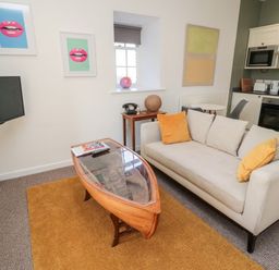 A living room with a beige sofa and yellow cushions a wooden boat-shaped coffee table and a small kitchen area at Cheviot View in Crookham near Cornhill-On-Tweed