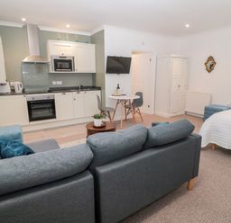 A studio apartment with a kitchen area a grey sofa a round dining table with two grey chairs a white wardrobe and a bed with a white cover at Flodden in Crookham near Cornhill-On-Tweed
